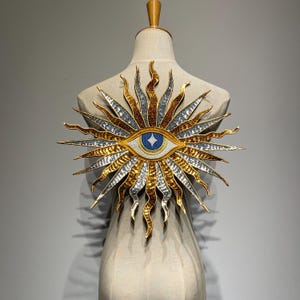 May include: A decorative sunburst-shaped brooch with an eye design. The brooch features gold, silver, and bronze metallic elements. The eye has a blue iris with a white diamond shape in the center. The brooch is displayed on a mannequin.