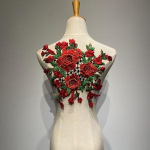 Può includere: Applicazione floreale rossa con ricamo di rose e foglie. L'applicazione, in rosso, verde e nero, è progettata per abbellire abiti o artigianato. Il design è esposto su un manichino.