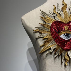 A red sequined heart with a white eye and eyelashes, surrounded by a gold and silver sunburst design.
