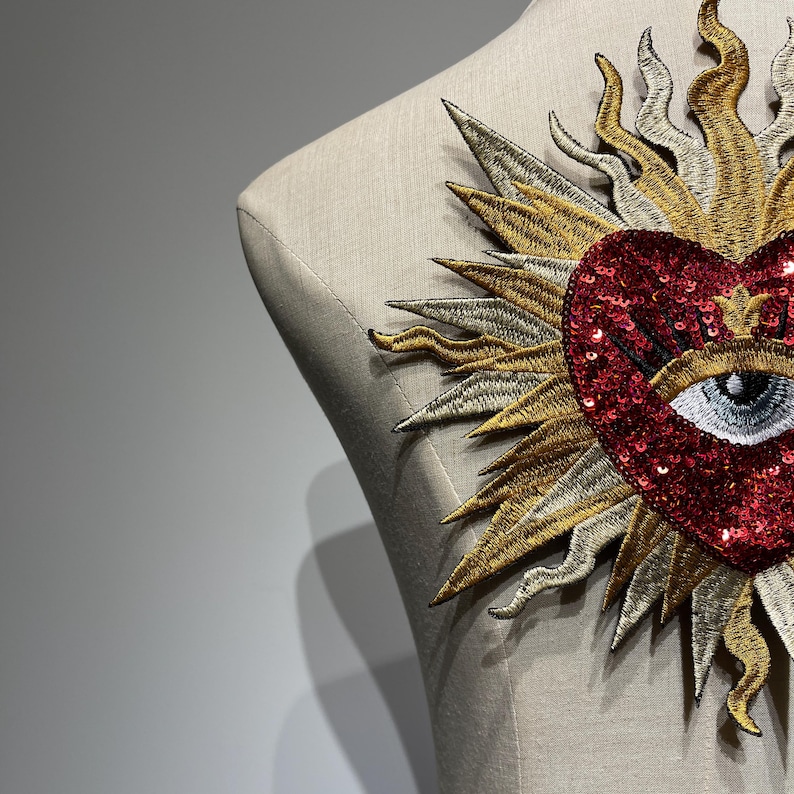 A red sequined heart with a white eye and eyelashes, surrounded by a gold and silver sunburst design.