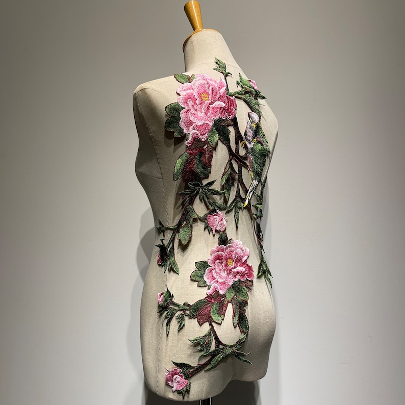 Pode incluir: Uma aplica&ccedil;&atilde;o floral rosa e verde com p&aacute;ssaros sobre um fundo branco. A aplica&ccedil;&atilde;o &eacute; feita de tecido bordado e &eacute; perfeita para adicionar um toque de eleg&acirc;ncia a qualquer projeto.