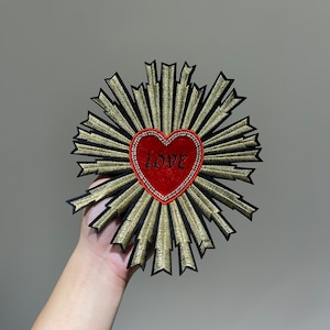 Può includere: Un cuore di feltro rosso con la parola "LOVE" ricamata su di esso, circondato da un design a stella dorata e nera.