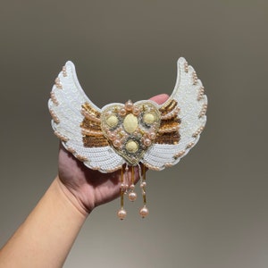 May include: A white beaded angel wing design with a heart-shaped center featuring gold and pink beads. The wings are embellished with sequins and the heart has a dangling chain of pink beads.