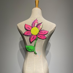 Puede incluir: Aplicación de flor grande con lentejuelas en rosa, amarillo y verde, exhibida en un maniquí. La flor tiene pétalos rosas con lentejuelas iridiscentes, un centro amarillo y un tallo y hoja verdes. Para ropa o accesorios.