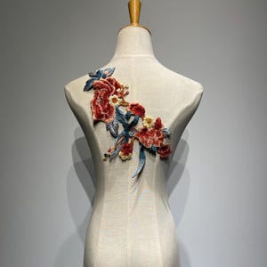 May include: Embroidered floral applique with red, yellow, and blue flowers and leaves. The applique is attached to a mannequin. The floral design is detailed and appears to be made of fabric.
