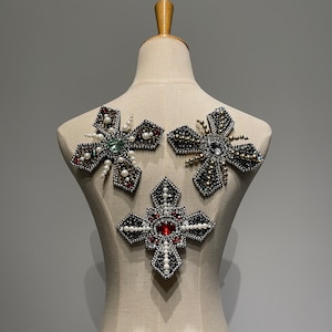 Puede incluir: Tres parches decorativos de tela en forma de cruz y dos estrellas. La cruz es plateada con una joya roja en el centro y perlas blancas alrededor de los bordes. Las estrellas son plateadas con una joya verde y una joya transparente en el centro y perlas blancas alrededor de los bordes.