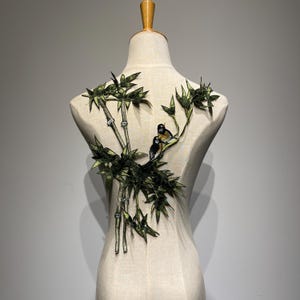 May include: Green embroidered applique featuring a bamboo branch with two birds perched on it. The applique is on a white fabric background.
