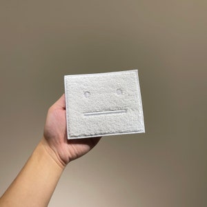 Op de afbeelding: Witte vierkante patch met een witte geborduurde neutrale gezichtsuitdrukking. De patch heeft een pluche textuur.