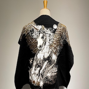 Puede incluir: Chal negro con una imagen grande y detallada de un unicornio blanco con alas y detalles dorados. El unicornio se encuentra sobre un fondo negro, con una luna llena en la esquina superior derecha. El chal se exhibe en un maniquí.