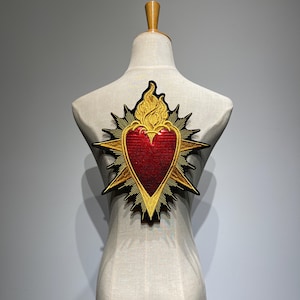 May include: Embroidered patch featuring a red heart with gold flames and a gold starburst design. The patch is attached to a mannequin. The heart has a textured appearance, and the flames are detailed with gold thread.