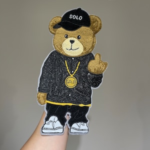 Peut inclure: Un ours en peluche brun portant une veste noire avec une chaîne en or et une casquette de baseball noire sur laquelle est inscrit "SOLO". L'ours porte également des baskets blanches.