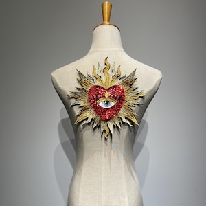 K&ouml;nnte beinhalten: Ein rotes Herz mit einem Auge in der Mitte, umgeben von einem goldenen Sonnenstrahl, ist auf eine Stoffpuppe gen&auml;ht.