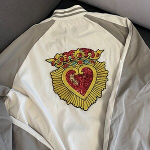 A white baseball jacket with a gold heart and crown design on the front. The heart is red with gold sequins and the crown is gold with red and blue accents.