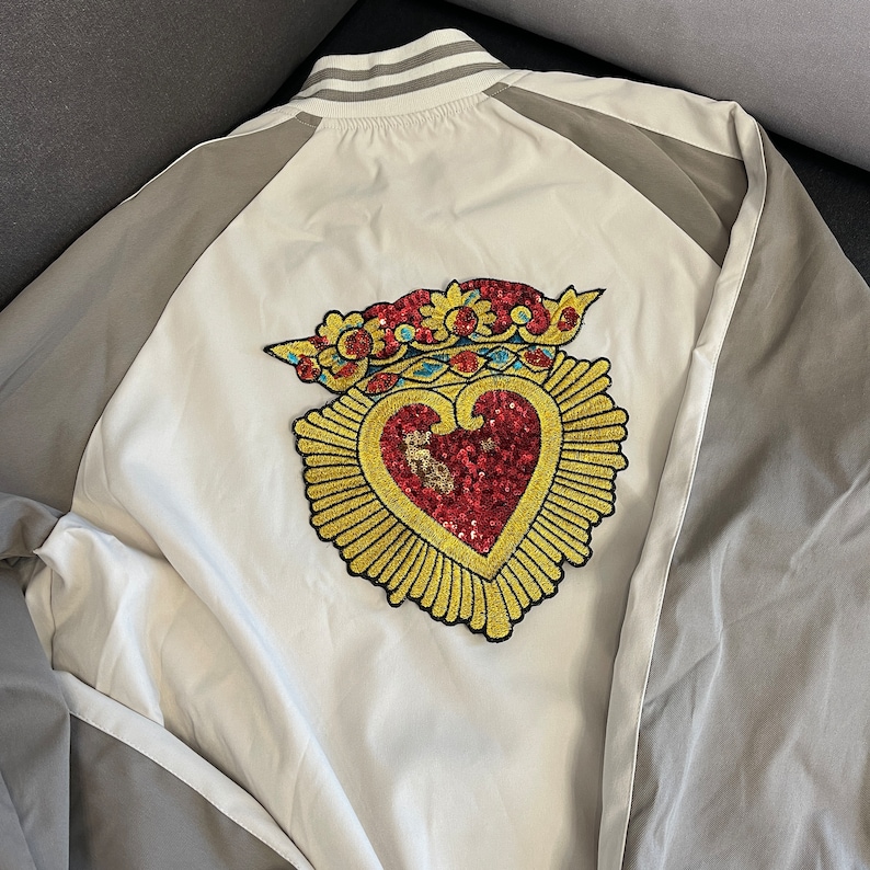 A white baseball jacket with a gold heart and crown design on the front. The heart is red with gold sequins and the crown is gold with red and blue accents.