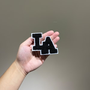 Può includere: Lettere "LA" in feltro nero con contorni bianchi su uno sfondo bianco. Le lettere sono tenute in una mano.