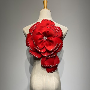 Könnte beinhalten: Eine große, leuchtend rote Stoffblume mit mehreren Schichten und einem perlenartigen Zentrum, verziert mit kleinen, funkelnden Strasssteinen an den Rändern. Die Blume wird auf einer Schaufensterpuppe präsentiert, was auf ihre Verwendung als Kleidungsstück-Verzierung hindeutet.