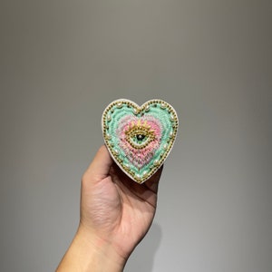Könnte beinhalten: Herzförmiger, bestickter Aufnäher mit einem zentralen Augendesign, einer Krone und Perlen. Der Aufnäher hat ein Pastellfarbschema aus Rosa, Grün und Gold mit einem weißen Rand.