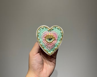 Può includere: Toppa ricamata a forma di cuore con un motivo a occhio centrale, una corona e perle. La toppa presenta una combinazione di colori pastello rosa, verde e oro, con un bordo bianco.