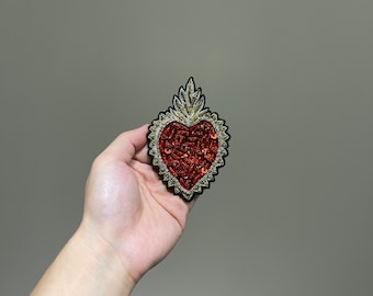 Puede incluir: Un parche en forma de corazón rojo con adornos dorados y plateados. El corazón está rodeado por un contorno negro y tiene un diseño en forma de llama en la parte superior.