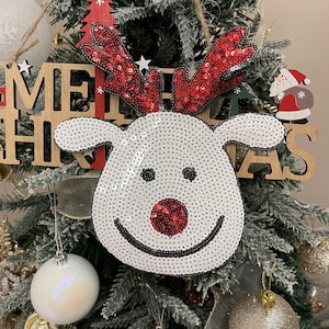 May include: A white reindeer ornament with red sequins for antlers and a red nose. The ornament is decorated with black sequins for eyes and a smile. The ornament is hanging on a Christmas tree with a wooden sign that says "Merry Christmas".