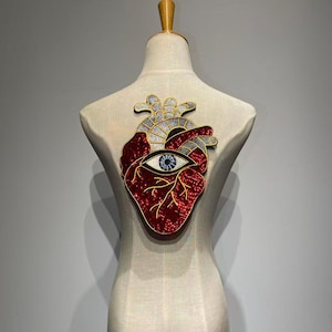 May include: A decorative heart-shaped applique with a red sequined design, featuring an eye and gold and silver accents. The applique is displayed on a mannequin, showcasing its intricate details and craftsmanship.