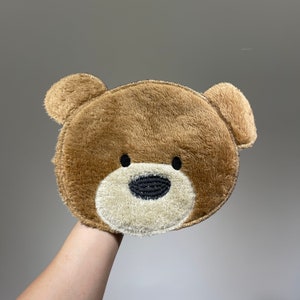 Puede incluir: Una cabeza de oso de peluche marrón con ojos y nariz bordados en negro. El oso es un diseño plano bidimensional.