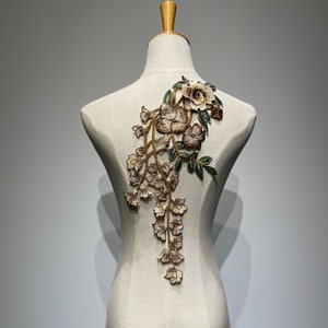 Op de afbeelding: Een delicate bloemenapplicatie met bruine en groene borduursels. Het ontwerp heeft een grote bloem aan de bovenkant met kleinere bloemen die langs de stengel naar beneden vallen.