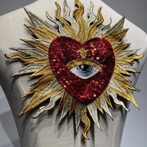 A red sequined heart with a black eye and eyelashes in the center, surrounded by a gold and silver embroidered sunburst design.