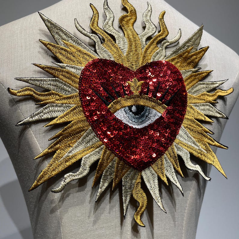 A red sequined heart with a black eye and eyelashes in the center, surrounded by a gold and silver embroidered sunburst design.
