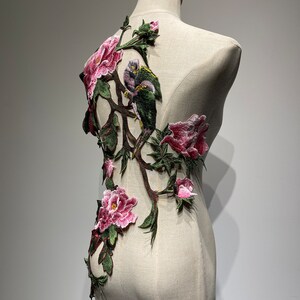 Pode incluir: Uma aplica&ccedil;&atilde;o bordada rosa e verde com p&aacute;ssaros e flores. A aplica&ccedil;&atilde;o est&aacute; sobre um fundo de tecido branco.