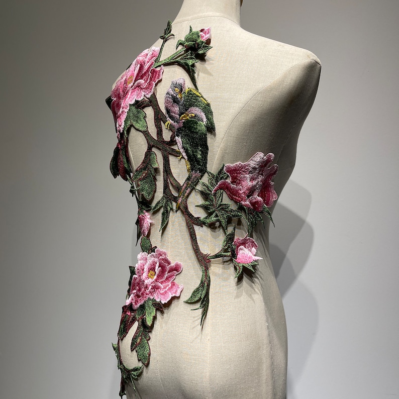 Pode incluir: Uma aplica&ccedil;&atilde;o bordada rosa e verde com p&aacute;ssaros e flores. A aplica&ccedil;&atilde;o est&aacute; sobre um fundo de tecido branco.
