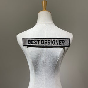 Puede incluir: Un parche rectangular negro con las palabras "BEST DESIGNER" bordadas en mayúsculas. El parche se muestra en un maniquí blanco. El parche tiene una textura similar a una malla.