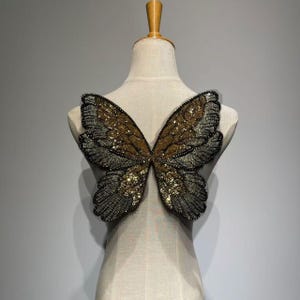 May include: A decorative butterfly applique with gold and black sequins. The butterfly wings are displayed on a mannequin. The wings have a detailed design with a black beaded outline and a gold sequined center.