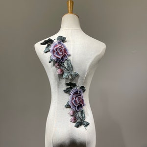 May include: Two embroidered purple and green rose appliques on a white fabric background.