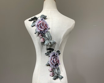 May include: Two embroidered purple and green rose appliques on a white fabric background.