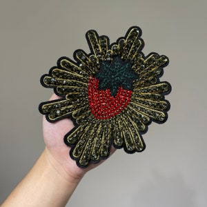 May include: A black fabric applique with a red and green embroidered strawberry design. The strawberry is surrounded by gold and black sequins.