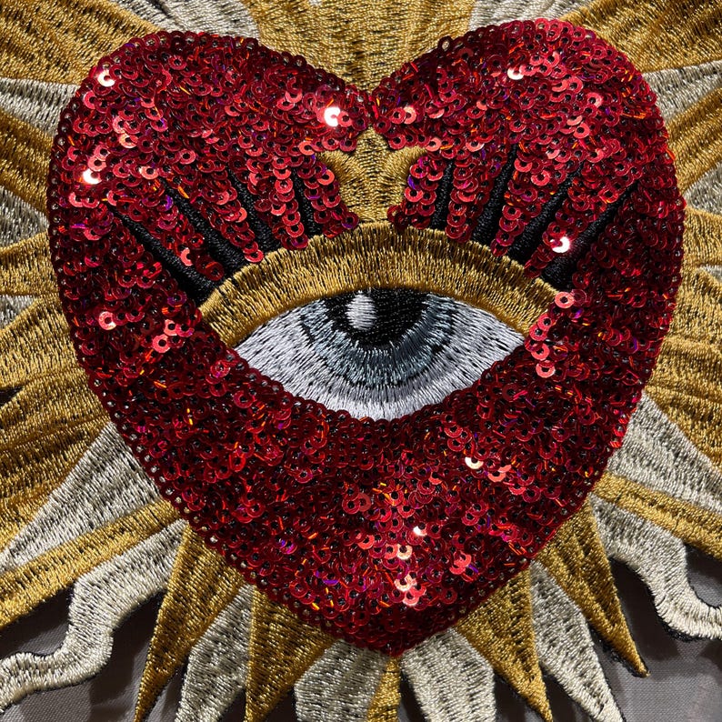 A red heart-shaped patch with a large embroidered eye in the center. The heart is covered in red sequins and the eye is outlined in black thread. The heart is surrounded by a golden embroidered sunburst pattern.