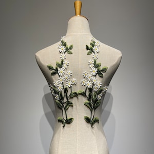 Puede incluir: Dos apliques de flores blancas con hojas verdes sobre un fondo de tela blanca. Los apliques tienen la forma de una enredadera floral con pequeñas flores blancas y hojas verdes.