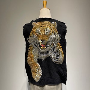 Puede incluir: Un chaleco negro sin mangas con un diseño de tigre hecho con adornos dorados y plateados. El tigre está de frente con la boca abierta y los dientes visibles.