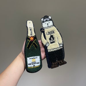 May include: Two embroidered patches, one of a champagne bottle with the text "Champagne Maison Roederer Vintage" and the other of a polar bear wearing a sweater with the text "Lodge & Snow".
