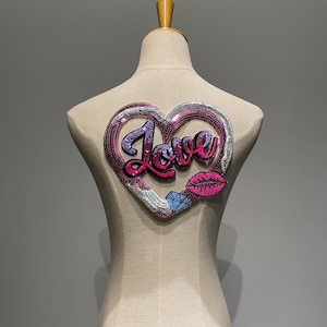 Peut inclure: Un patch en forme de cœur en sequins rose, blanc et bleu avec le mot "Love" en lettres roses et un motif de lèvres roses.