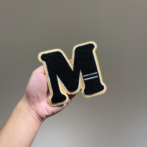 Peut inclure: Une lettre M en feutre noir avec des coutures blanches sur un fond en feutre beige. La lettre est légèrement surélevée par rapport au fond.