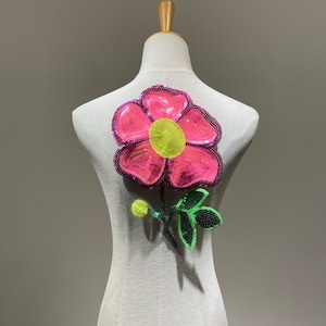 May include: A pink and green sequined flower applique with a yellow centre. The flower is on a white background.