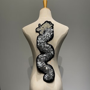 May include: A silver and gold embroidered dragon applique with a black backing. The dragon is curled in a serpentine shape.