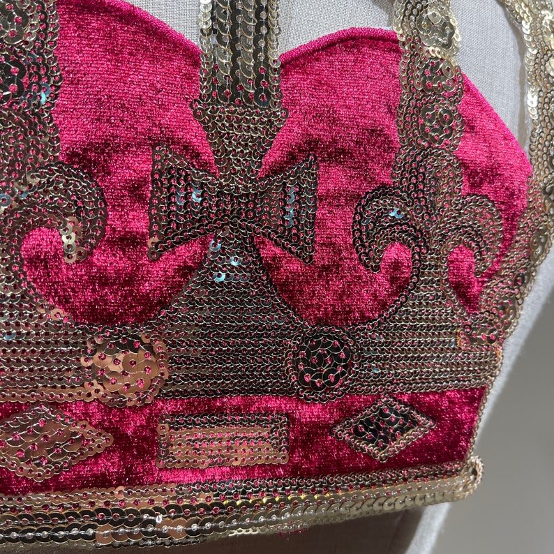 May include: A close-up of a red velvet crown with gold sequin embellishments. The crown has a pointed top and a rounded base.