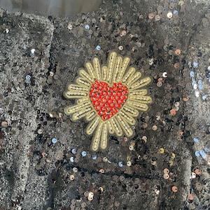 May include: Black fabric covered in sequins with a gold and red heart-shaped embellishment. The heart is surrounded by a gold floral design.