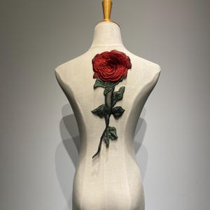 Könnte beinhalten: Eine rote gestickte Rose mit grünen Blättern und Stiel auf einem weißen Stoffhintergrund.