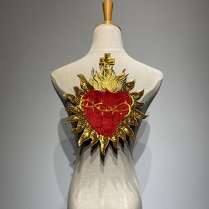 Puede incluir: Parche bordado en oro y rojo que representa un corazón con una corona de espinas, llamas y una cruz. El parche se exhibe en un maniquí, mostrando los detalles intrincados y los colores vibrantes del símbolo religioso.