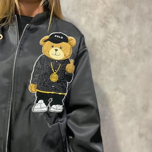 Puede incluir: Chaqueta bomber de satén negro con un gran parche bordado de un oso de peluche marrón que lleva un gorro negro que dice "SOLO" y una cadena de oro con un colgante "GOLD". El oso está haciendo un pulgar hacia arriba.