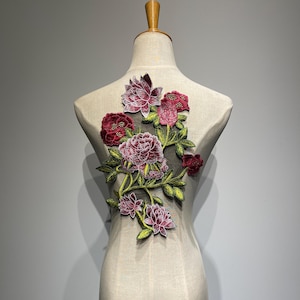 Könnte beinhalten: Gestickte Blumenapplikation mit burgunderroten, rosa und weißen Blumen und grünen Blättern. Die Applikation wird auf einer Schaufensterpuppe präsentiert und zeigt ihre Verwendung zur Dekoration von Kleidungsstücken. Das Design ist aufwendig.